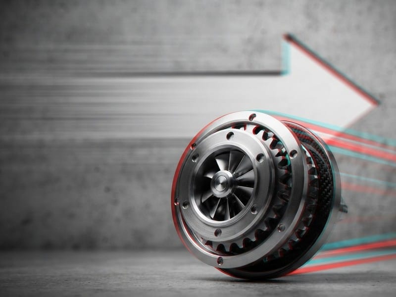 A metallic mechanical turbine component rolling forward against a textured concrete wall with a large directional arrow and motion blur lines.